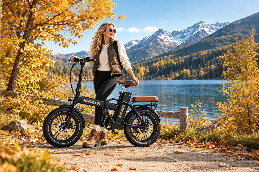 Blonde woman standing with black KOOLUX foldable fat tire electric bike beside alpine lake, golden fall trees and snow mountain backdrop, outdoor autumn cycling