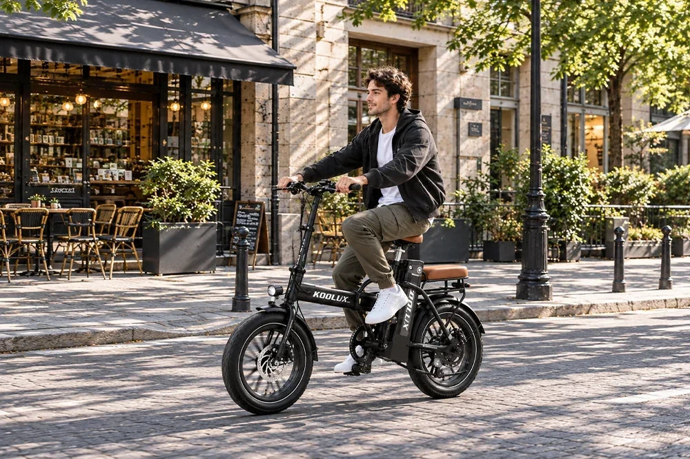 Casual young man riding black KOOLUX foldable fat tire electric bike on cobblestone street in front of Parisian-style sidewalk café, dappled tree shade, urban weekend ebike lifestyle