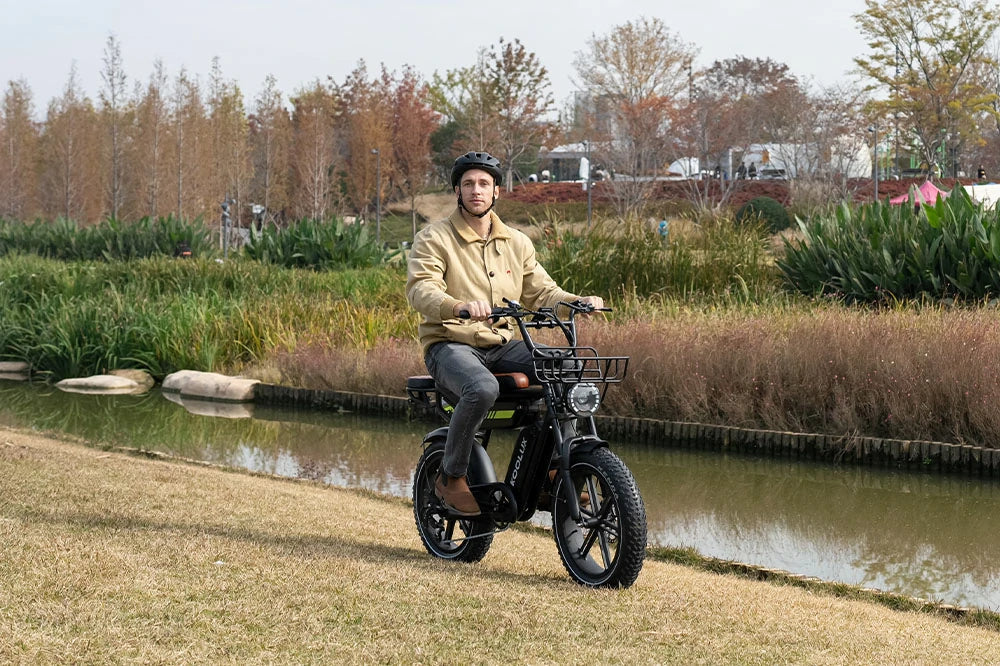 Lifestyle photo of a man wearing a helmet riding the KOOLUX X9 Pro electric bike with fat tires in a riverside urban park, showcasing the bike's black frame, front basket, and outdoor adventure-ready design
