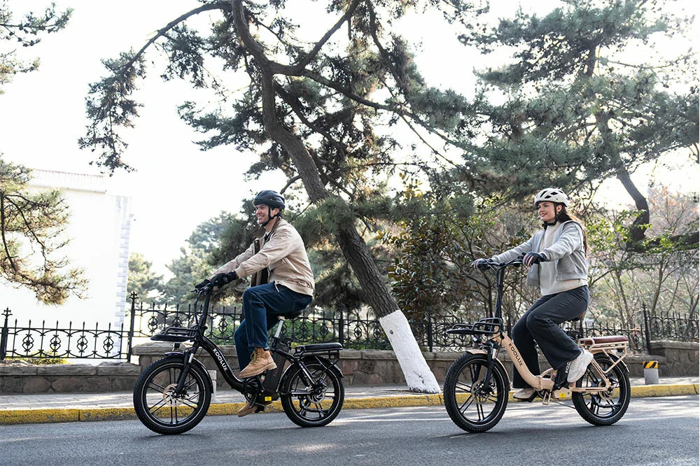Couple riding black and beige KOOLUX foldable fat tire electric bikes side by side on quiet city street, pine tree background, casual outdoor travel lifestyle photo